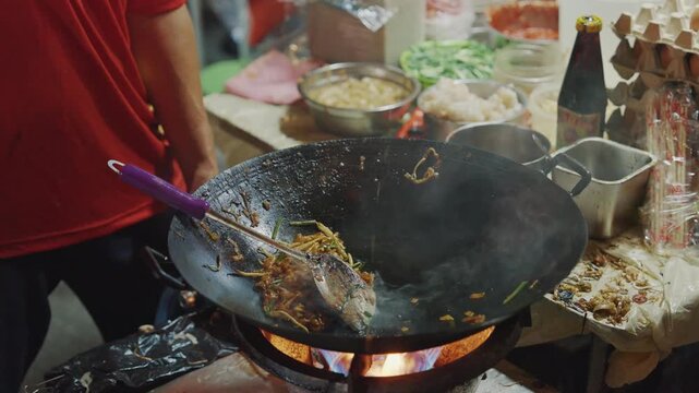 A close-up action shot of a street food hawker stir-frying Char Koay Teow (flat rice noodles) with prawns and bean sprouts over a roaring blue gas flame in a large iron wok.