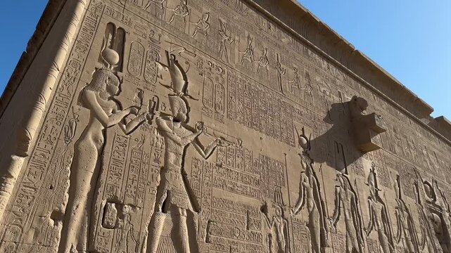 Cinematic panning shot of the exterior stone wall of the Temple of Hathor in Dendera featuring detailed ancient Egyptian reliefs of pharaohs and gods under bright daylight