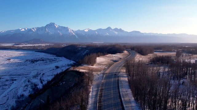 Low drone flight over Glenn Highway near Palmer, AK heading toward dramatic snow-capped peaks. Frozen river bed flanks left, bare winter trees line both sides, hazy mountain range fills horizon. DJI A