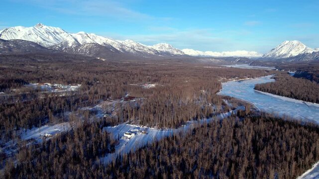 High altitude drone shot over Matanuska Valley near Palmer, AK. Winding frozen river curves right through dense forest, rural homesteads visible below, snow-capped peaks encircle entire valley. DJI Ai