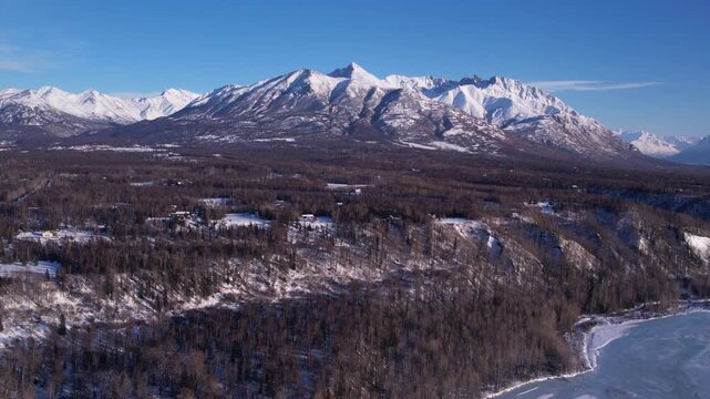 Medium altitude straight drone flight over snow-dusted forest and rural homesteads near Palmer, AK. Massive Chugach peaks dominate horizon, frozen river edge visible lower right. DJI Air 2S 4K 60fps.
