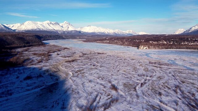 Medium altitude drone flight over snow-dusted Matanuska River gravel bars near Palmer, AK. Partially frozen channel curves left, rocky bluff mid-frame, full snow-capped range stretches across entire h