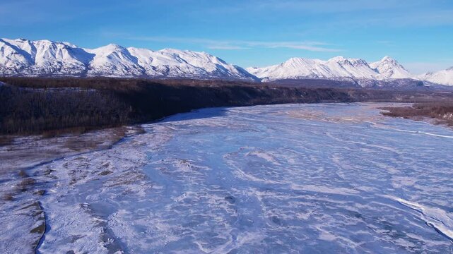 Drone ascending straight up over frozen Matanuska River near Palmer, AK. Textured ice expanse widens as altitude increases, revealing full mountain range and forested bluff stretching across horizon.