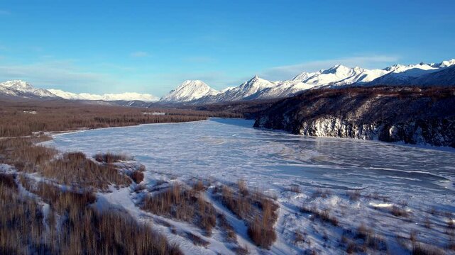 Medium altitude drone flight toward frozen Matanuska River and snow-capped peaks near Palmer, AK. Dry winter grass flanks left bank, rocky snow-covered bluff dominates right, mountain range fills hori