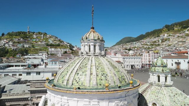 Aerial drone circles ornate green tile dome of Iglesia Compania de Jesus in Quito with El Panecillo hill and colonial cityscape behind