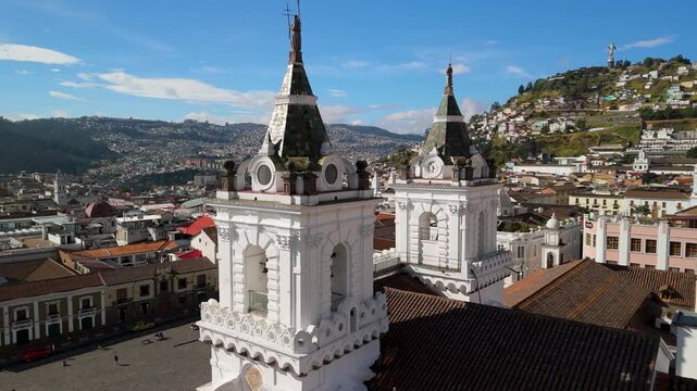 Drone orbits close around twin white baroque bell towers of Convento San Francisco in Quito with El Panecillo Virgin statue and cityscape behind