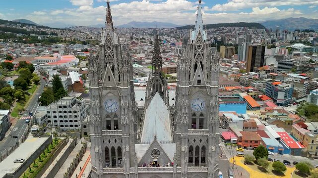 Drone flies between Gothic clock towers of Basilica del Voto Nacional in Quito revealing spires, nave roof, cloister and sweeping city panorama