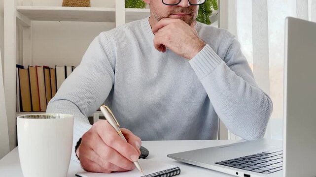 Man in a blue sweater works on a laptop at a clean white desk, surrounded by books, a cup of coffee, and a green plant. Remote work and the comfort of working from home. Perfect for a digital nomad