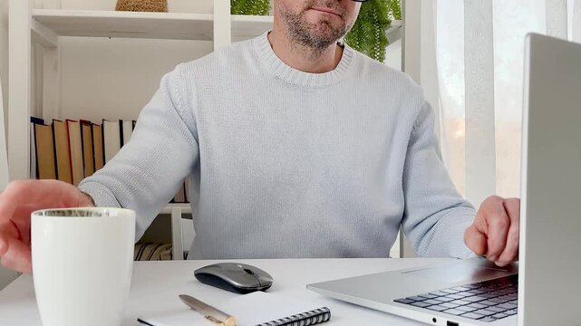 Man in a blue sweater works on a laptop at a clean white desk, surrounded by books, a cup of coffee, and a green plant. Remote work and the comfort of working from home. Perfect for a digital nomad