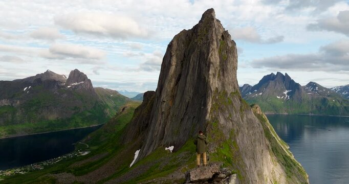 Cinematic aerial of a explorer beside Segla on Senja Island, Norway, overlooking a fjord, jagged peaks and reflective water beneath the midnight sun in northern Scandinavia