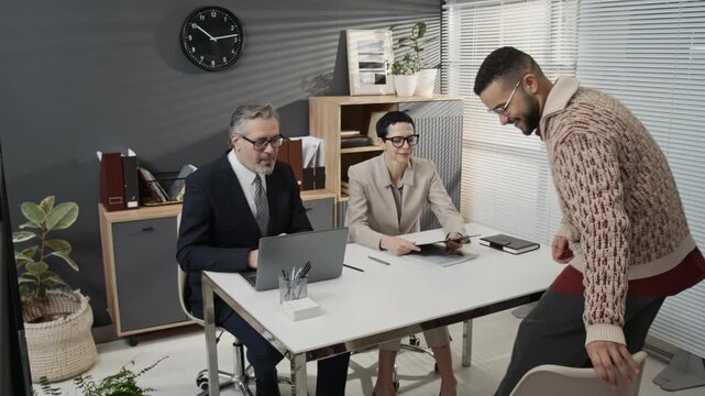 Gen Z jobseeker coming to office desk and handing printed resume to short-haired female HR manager, middle-aged businessman inviting him to sit down in front of them