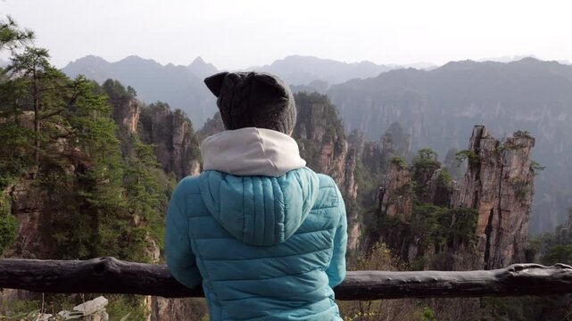 Slow motion parallax shot from behind female traveler standing on observation deck, admiring majestic quartz sandstone pillars of Zhangjiajie National Forest Park during soft light at golden hour.