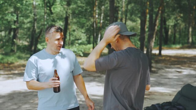 Hikers relaxing with drinks in forested area. Casual friends converse while resting near tranquil water. Two individuals enjoy beer and conversation amidst natural surroundings and scenic views