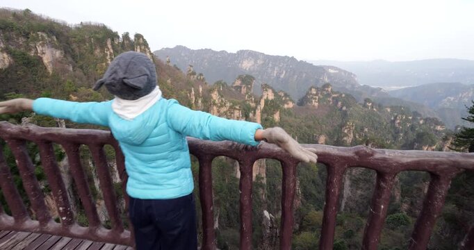 Tourist woman standing on wooden observation deck with her arms outstretched, enjoying breathtaking view of quartz sandstone pillars of Wulingyuan National Park in China, camera move forward
