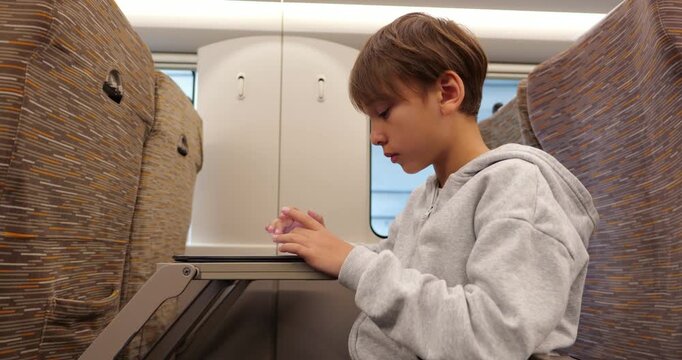 Concentrated boy wearing grey hoodie sitting in passenger seat on high speed train, deeply engaged in playing puzzle game on digital tablet during his journey. Side view, no other people visible