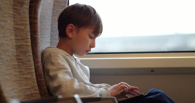 Concentrated young boy sits by window inside modern high-speed train, fully engrossed in digital tablet. He plays games or types, outdoor scenery flashes by in high-speed motion blur through glass