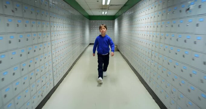 Energetic boy in blue hoodie walking confidently toward tracking camera as it glides backward through symmetrical hallway lined with rows of metal P.O. boxes. Wide-angle dynamic shot