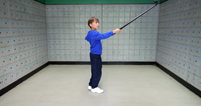 Young boy turn around with action camera on selfie stick to record 360 video of himself surrounded with symmetrical rows of small PO boxes in three walls.