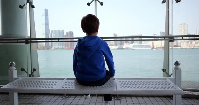 Little boy sitting on bench in front of large window, dangling his legs while contemplating urban Kowloon landscape across water. Moment of quiet wonder while waiting for ferry in Hong Kong