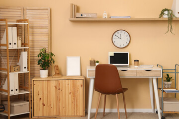 Fototapeta premium Interior of stylish home office with laptop, workplace and clock hanging on beige wall