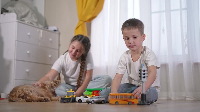 Girl strokes fluffy puppy on warm floor as boy moves toy truck. Kids and puppy enjoy shared time with vehicles and blocks. Sibling bonds near window. Girl, boy, puppy, toy frame peaceful moment.