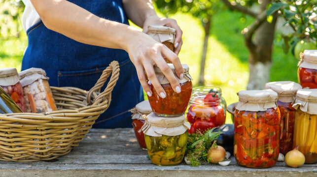 Woman putting jars of preserved vegetables into wicker basket on wooden table