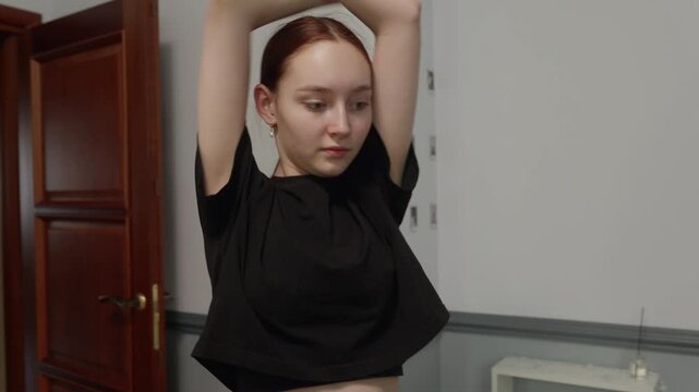 A young woman with red hair in a black crop top warms up and begins practicing a contemporary or modern dance routine in her bedroom. She stretches and moves with fluid, expressive motions.