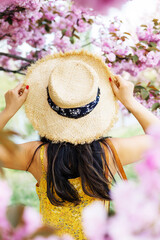 A high-angle, rear-view photograph of a woman wearing a wide-brimmed straw hat and a yellow floral dress, looking into a vibrant canopy of pink cherry blossoms. The woman’s hands are gracefully holdin © Mykola