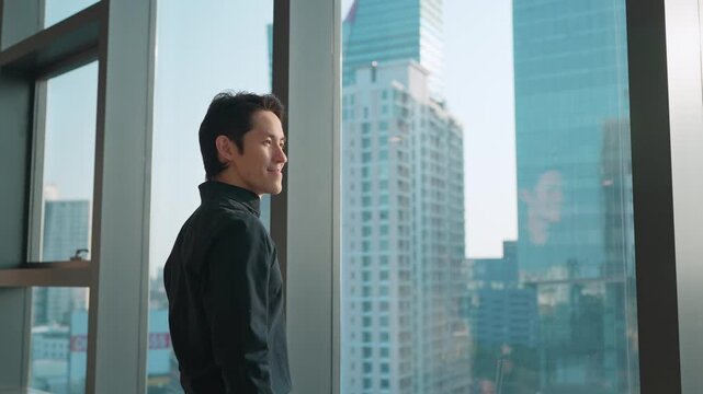 Young Asian businessman looking out the window at a modern city skyline from a high-rise office, symbolizing confidence, ambition, leadership, and future business vision