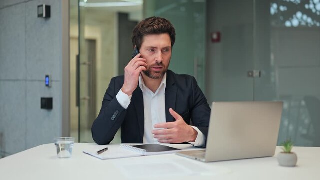 Focused businessperson on phone call in modern office. Conveying serious discussion and active problem-solving while consulting laptop. Expresses concentration and commitment to tasks.