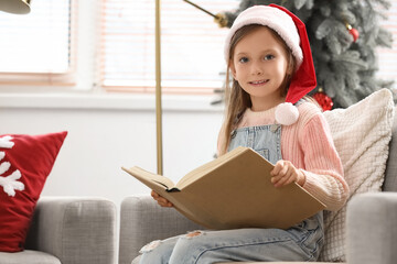 Naklejka premium Cute little girl in Santa hat reading Christmas story on sofa at home