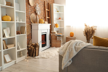 Naklejka premium Interior of living room with armchair, wicker basket, shelving units and autumn home decor on fireplace near brick wall