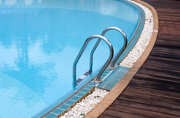 Swimming pool with stainless steel ladder and clear blue water. Luxury resort outdoor poolside with wooden deck and white stones.Summer vacation background for travel,Hotel leisure relaxation concept
