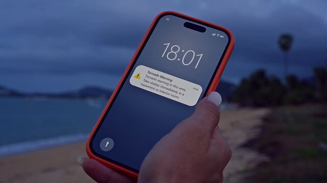 Woman Checking Smartphone With Tornado Warning Notification While Standing on Windy Beach Under Dark Storm Clouds Zoom Shot