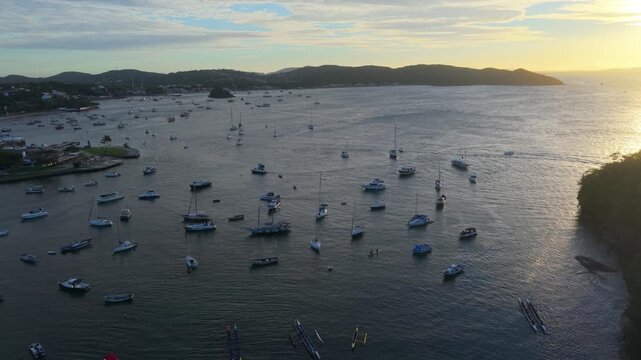 Breathtaking drone footage of sailboats and yachts anchored in the calm waters of Praia dos Ossos in Buzios, Brazil, during a beautiful golden hour.
