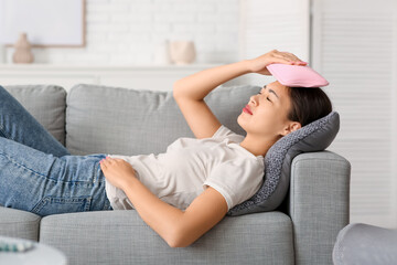 Obraz premium Young Asian woman warming her head with hot water bottle on sofa in living room