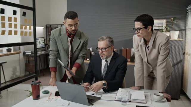 Zoom out shot of three diverse coworkers discussing business project in office, middle-aged man sitting in front of laptop, gen Z guy showing data on tablet, elegant mature woman watching them