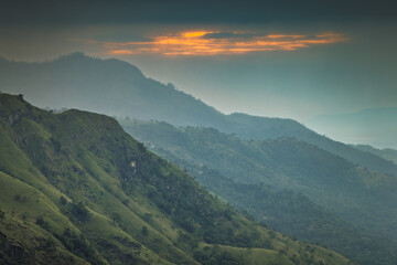Fototapeta premium Dramatic sunrise Glow Over Lush Green Mountain Peaks and Valleys with Hazy Atmosphere and Vibrant Sky Colors. Mountain landscape near Ella in Sri Lanka.