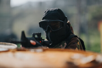 Fototapeta premium Masked paintball player in camouflage aims a marker with a scope. The scene shows a tactical shooter ready for action.