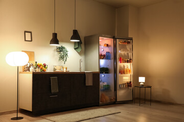 Open fridge full of different fresh products with lamps and utensils on counter near beige wall in...