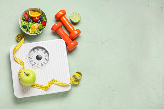 Weight scales with measuring tape, dumbbells and healthy food on grunge green background. Slimming concept