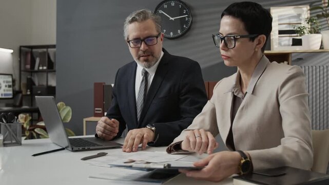 Two middle-aged colleagues in formalwear comparing resumes of job candidates when making decision sitting at work table in office