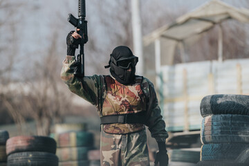 Fototapeta premium A camouflaged paintball player raises a rifle in a tactical stance, outdoors among tires and obstacles.