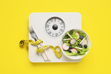 Weight scales with measuring tape, cutlery and bowl of fresh salad on yellow background. Slimming...