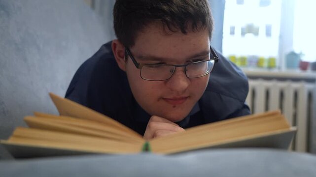 Reading book and doing homework. A teenage boy with glasses reads a book lying on the couch during the day.
