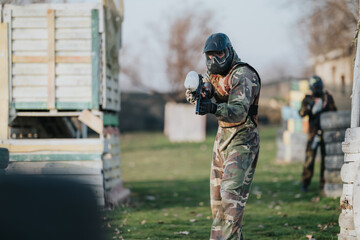 Fototapeta premium A camouflaged paintball player in a protective mask aims a marker on a grassy field. Another player hides behind barriers in the background during a daytime game.