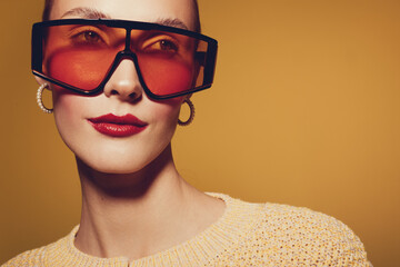 Stylish woman with oversized orange sunglasses and gold hoop earrings poses against a warm yellow background. Fashion portrait with bold accessories and confident expression. © SHOTPRIME STUDIO