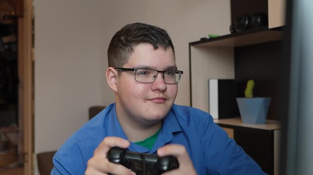 Teenager playing video games at home. A teenager in glasses sits at a computer holding a joystick, his face focused but sad, expressing disappointment after losing a video game.