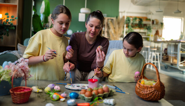 Mother and her two daughters engaging in a creative activity, decorating easter eggs with colorful paints at a table in a bright, modern home, enjoying holiday traditions