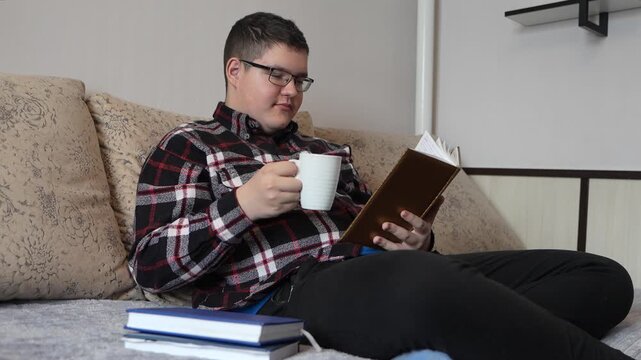 A smart teenager in glasses sits with a book and a coffee mug in the morning, reading a novel or textbook for school preparation.